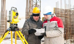 Two construction surveyors using a total station and reviewing plans on an urban construction site, demonstrating advanced construction surveying techniques.