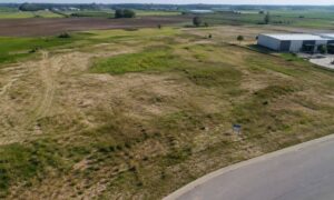 Aerial view of a vacant grassy lot with a curved road in the foreground and a white industrial building to the right.