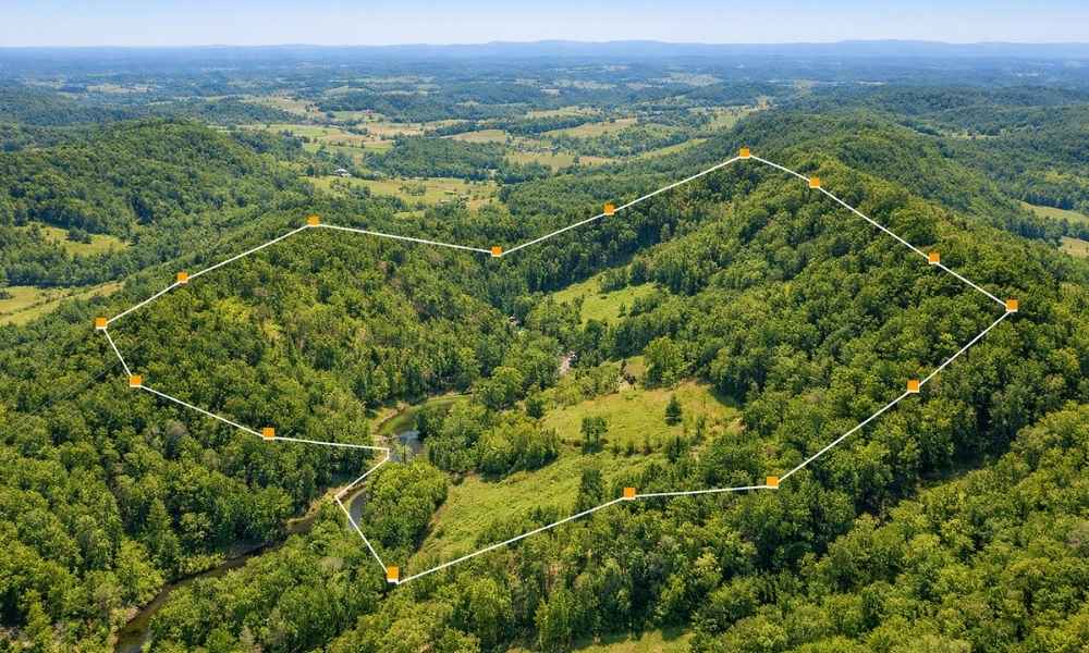 Aerial view of an irregular-shaped wooded property with survey boundary lines over rolling hills and a creek, illustrating factors that can increase property survey costs